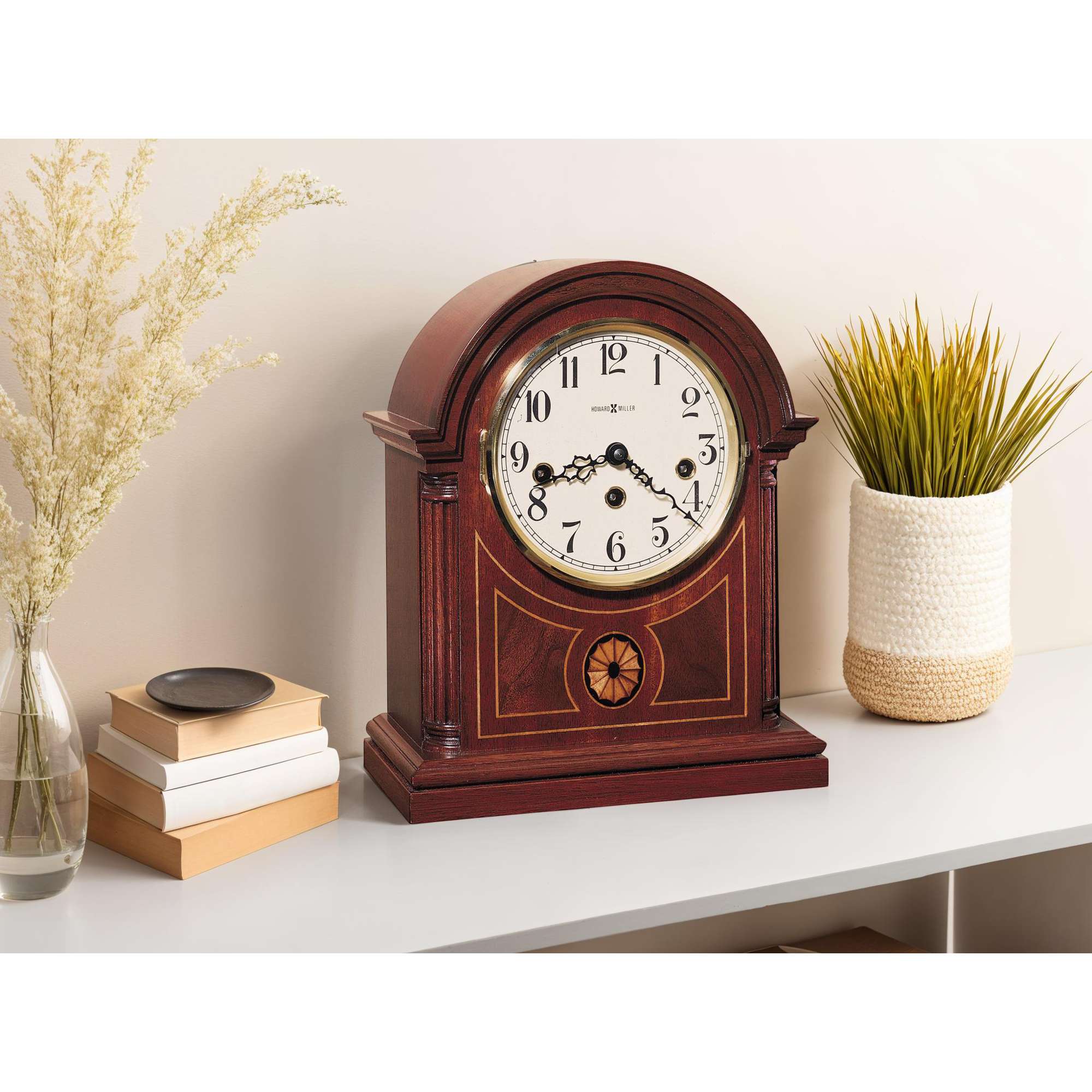 A classic wooden mantel clock with a white face and black numbers, placed on a modern shelf alongside books and a decorative plant.