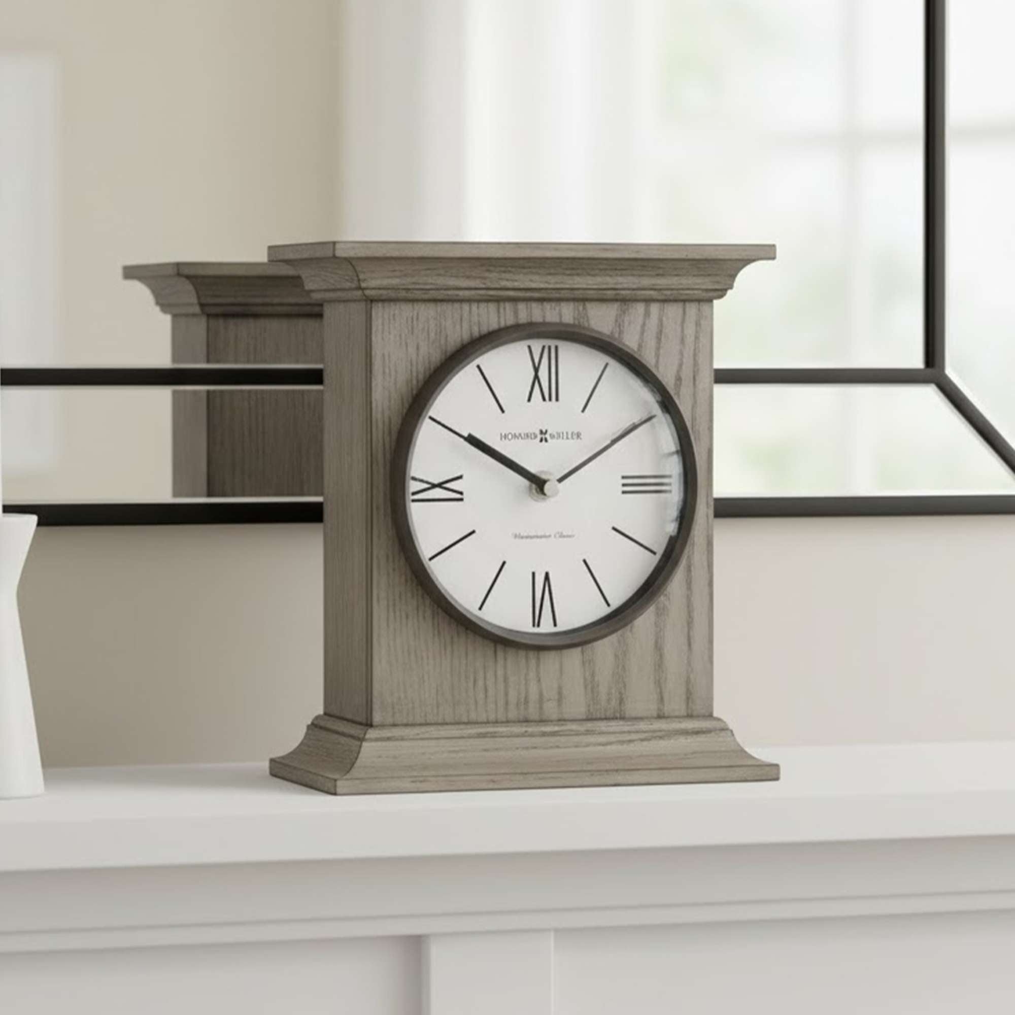 A round wall clock with Roman numerals displayed on a wooden stand, placed on a shelf near a mirror.