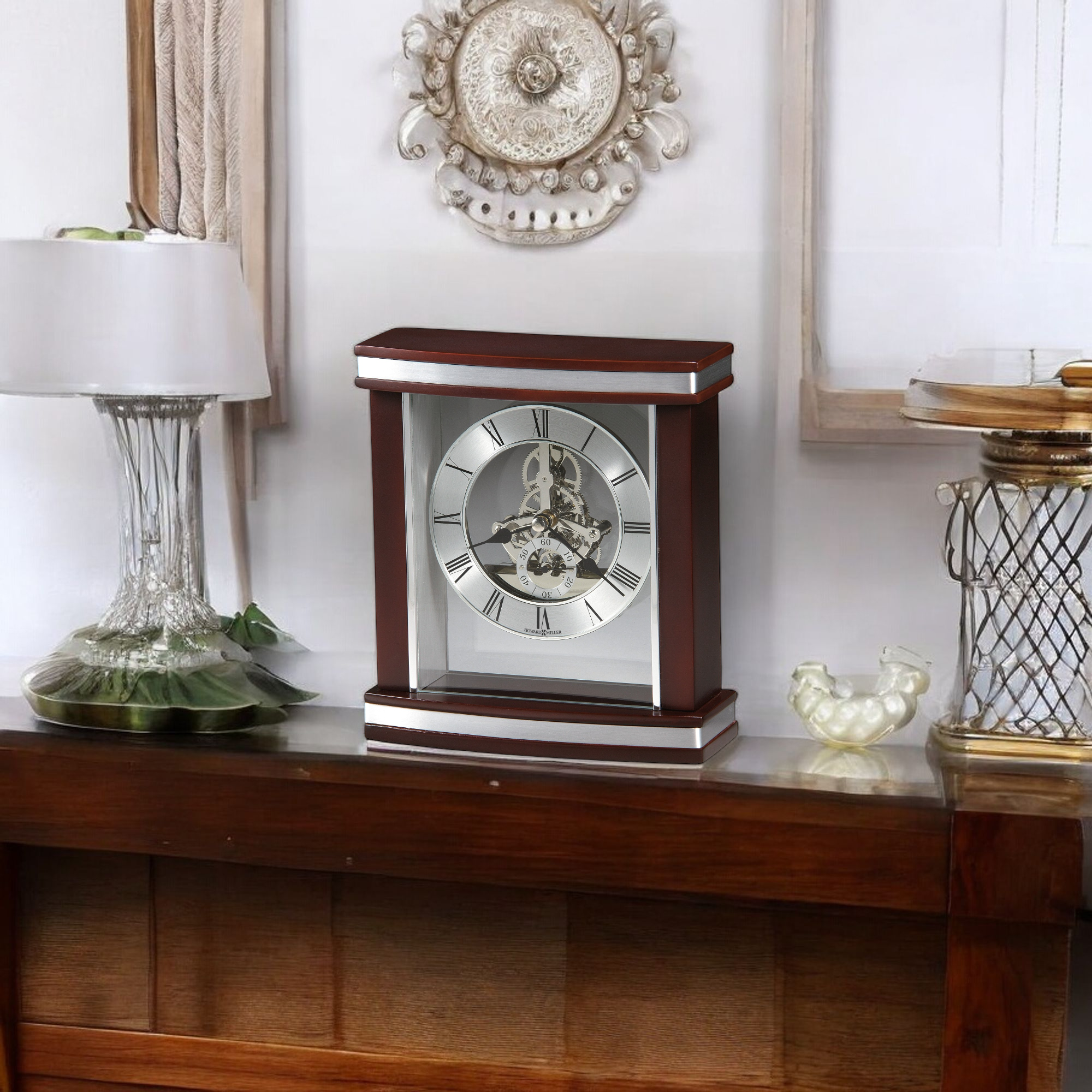 A decorative mantel clock on a wooden shelf with a lamp and other decorative items.
