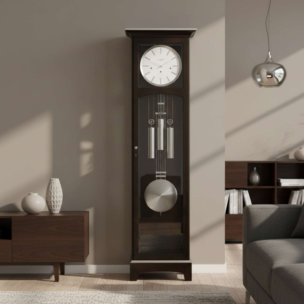 A tall wooden grandfather clock with a white face, pendulum, and polished metal chimes, set against a modern living room backdrop.