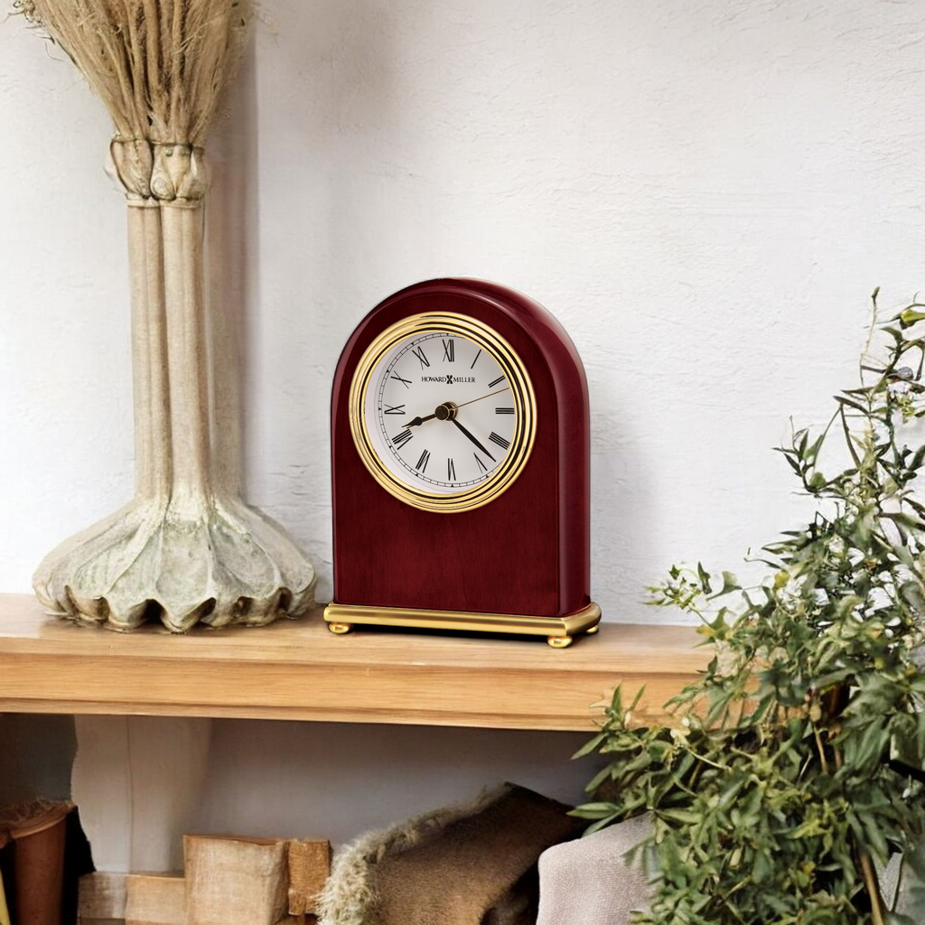A wooden clock with a classic design placed on a wooden shelf next to greenery.