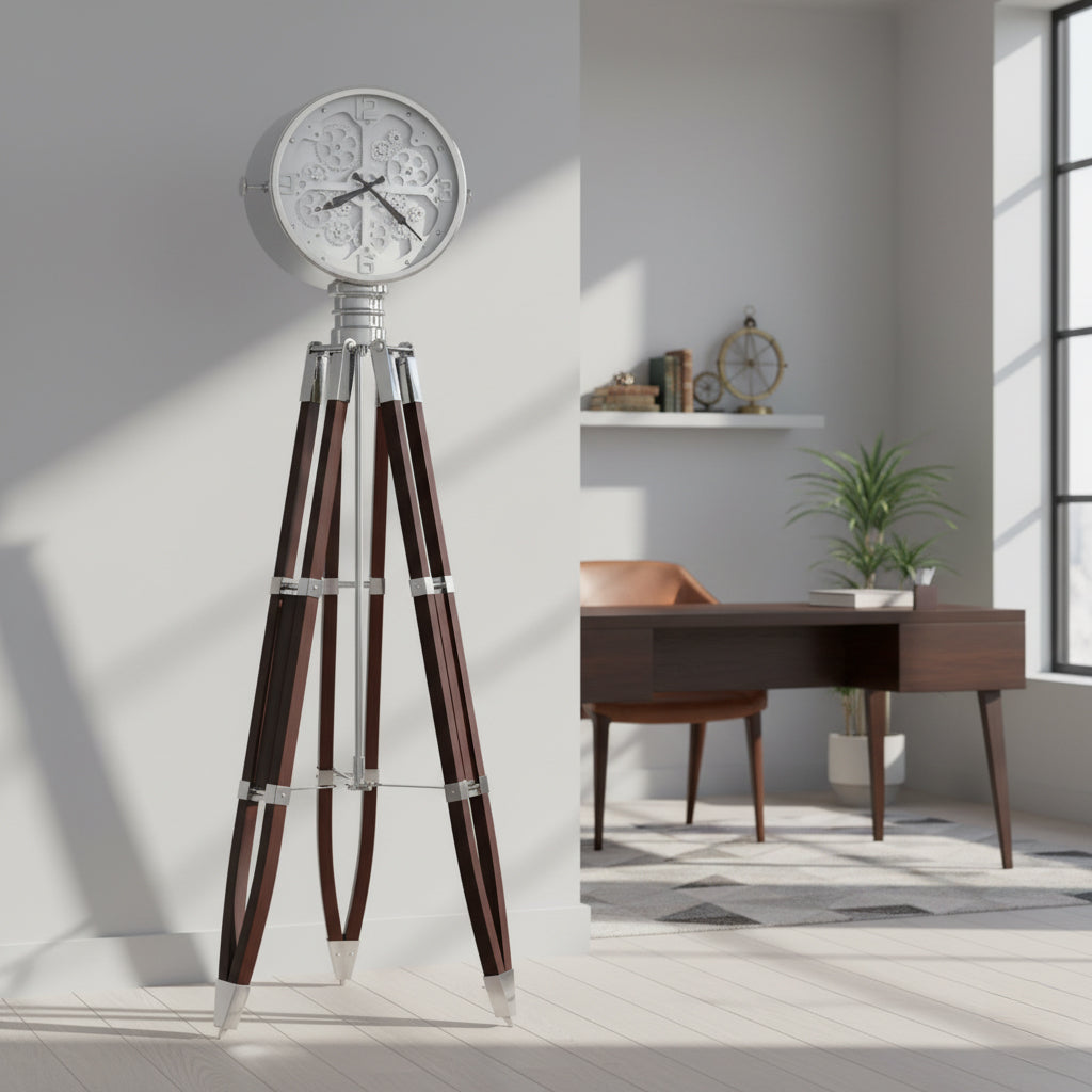 A modern floor clock with a silver clock face and wooden tripod legs in a bright room with a desk and potted plant.