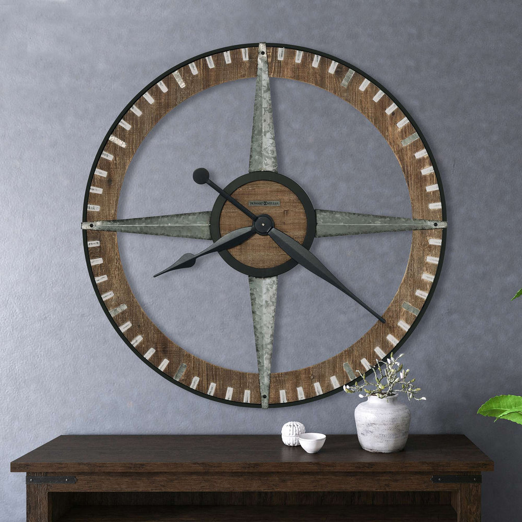 A large decorative wall clock resembling a compass mounted on a grey wall, with a wooden shelf underneath.