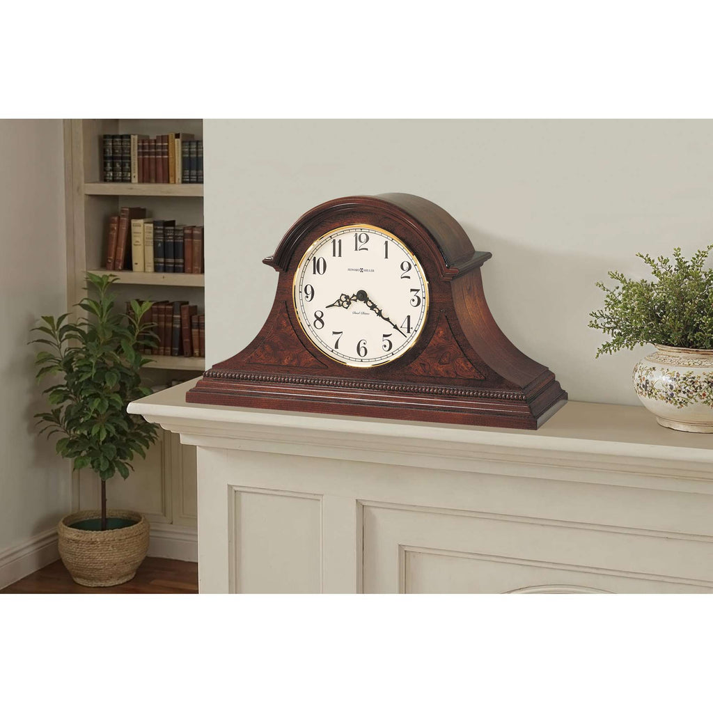 A vintage-style clock resting on a mantelpiece with a plant and bookshelf in the background.