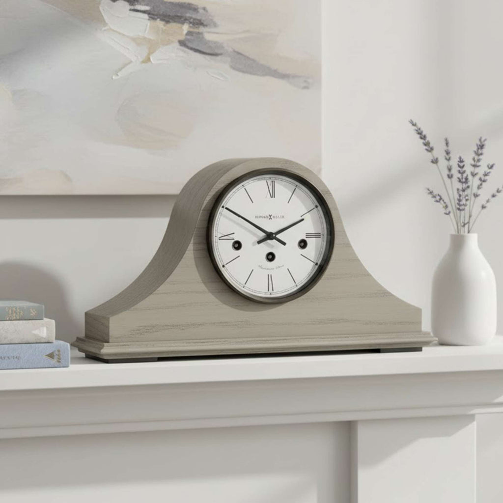 A decorative mantel clock with a subtle design, placed on a shelf with a vase and books.