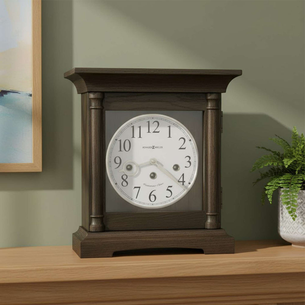 A classic wooden tabletop clock with a round face and Roman numerals, placed on a wooden surface next to a potted plant.