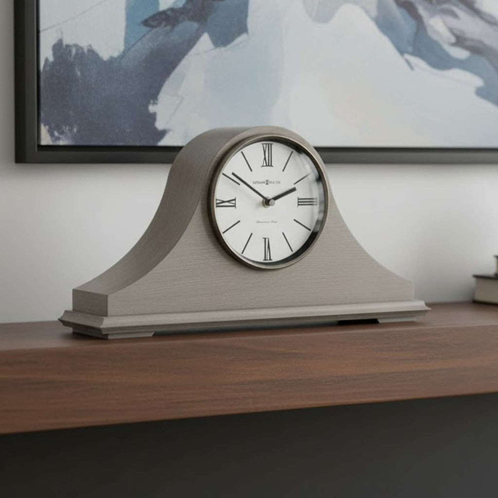 A stylish gray mantel clock on a wooden shelf against a modern abstract painting.