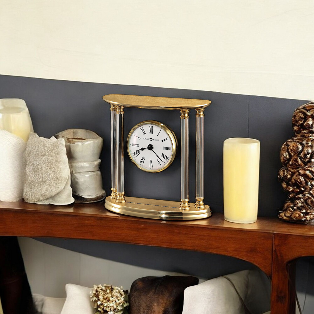 A decorative gold clock with glass columns on a wooden shelf, surrounded by candles and decorative items.