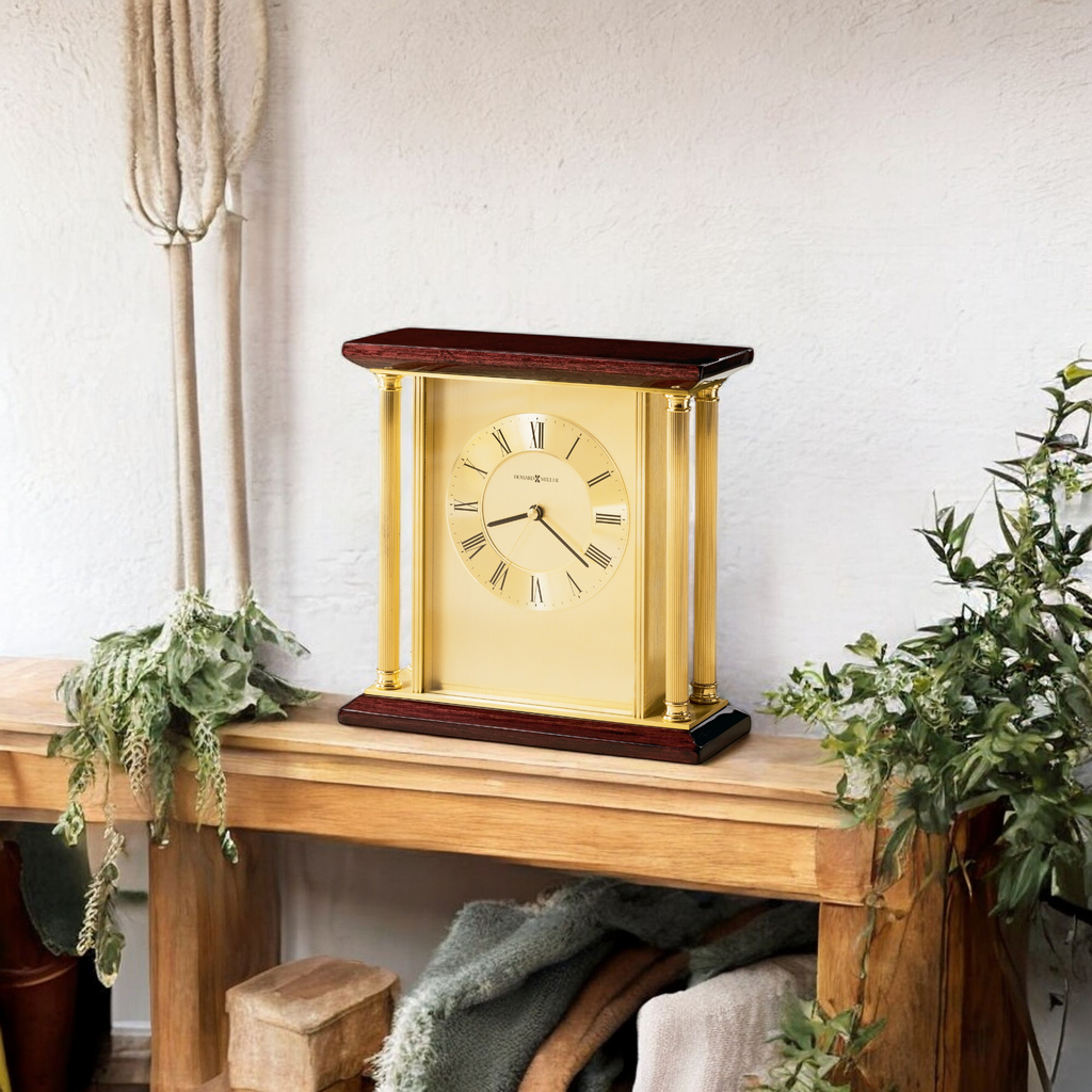 A classic gold and wood mantel clock set on a wooden table with greenery in a cozy interior.