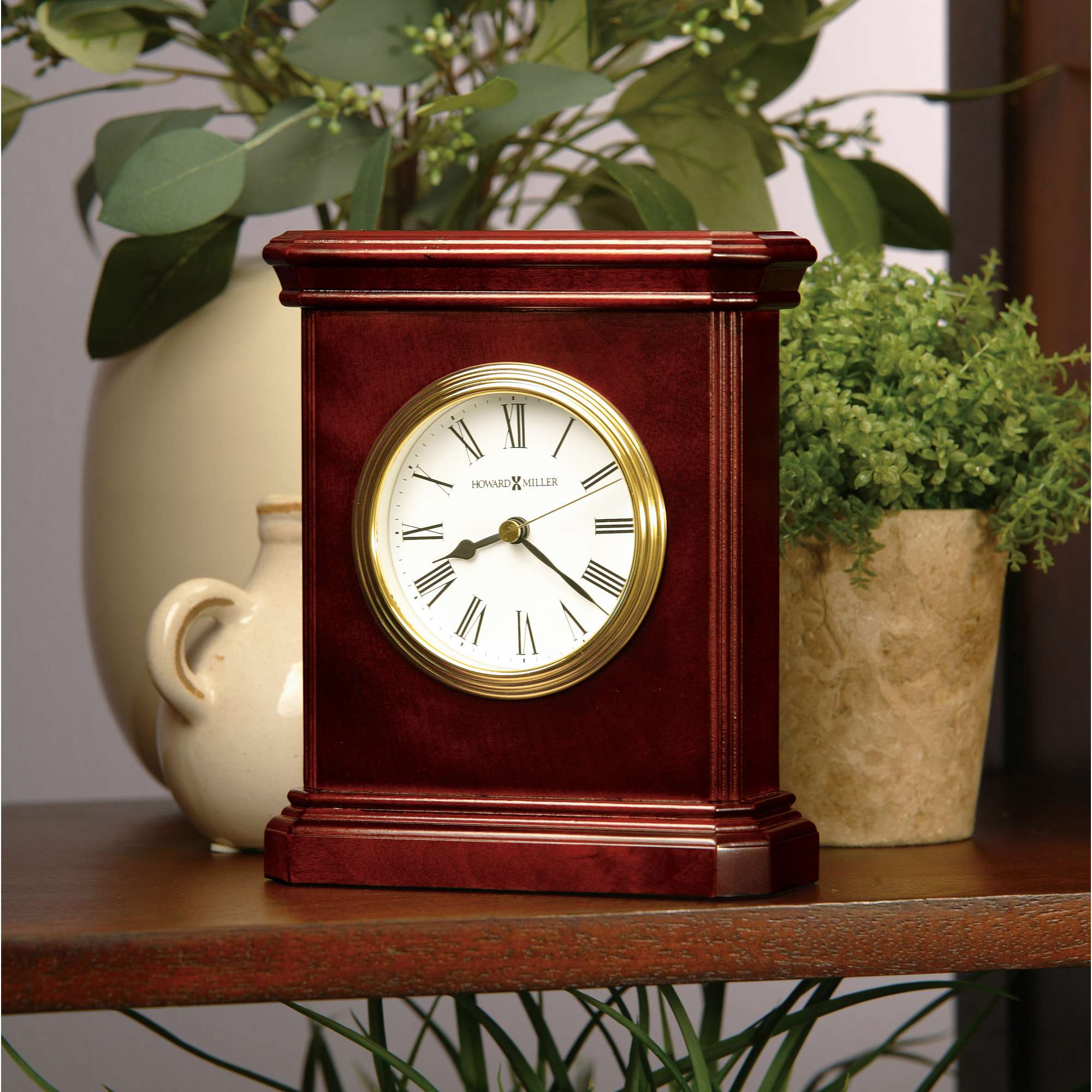 Elegant wooden clock with a round white face and gold detailing on a shelf with greenery and decorative vase.