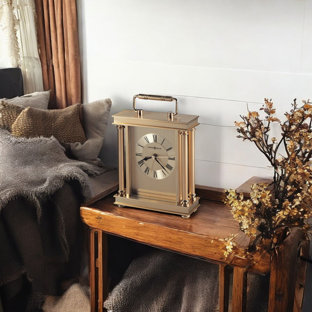 A vintage-style gold clock on a wooden table next to a cozy couch with blankets and a vase of dried flowers.
