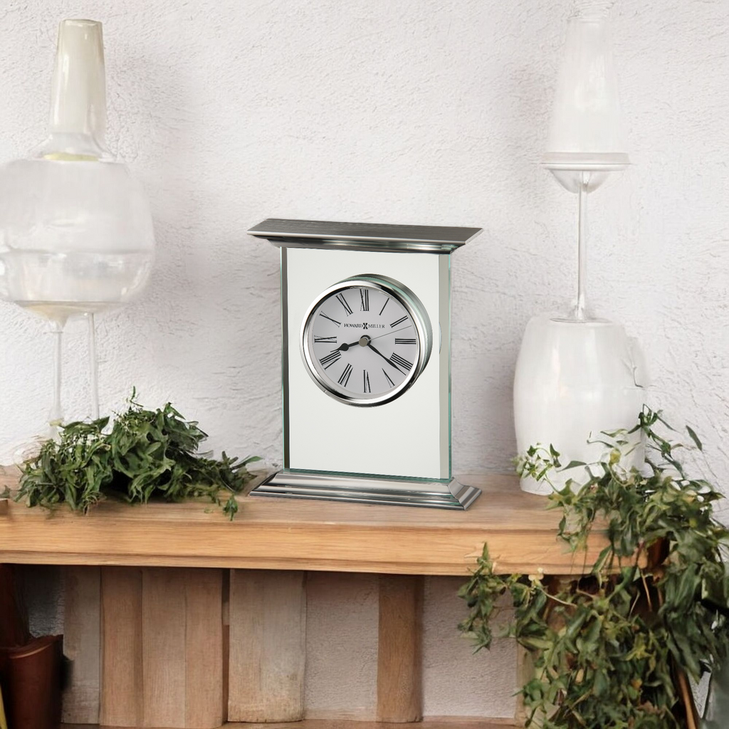 A stylish clock on a wooden shelf surrounded by greenery and decorative glassware.