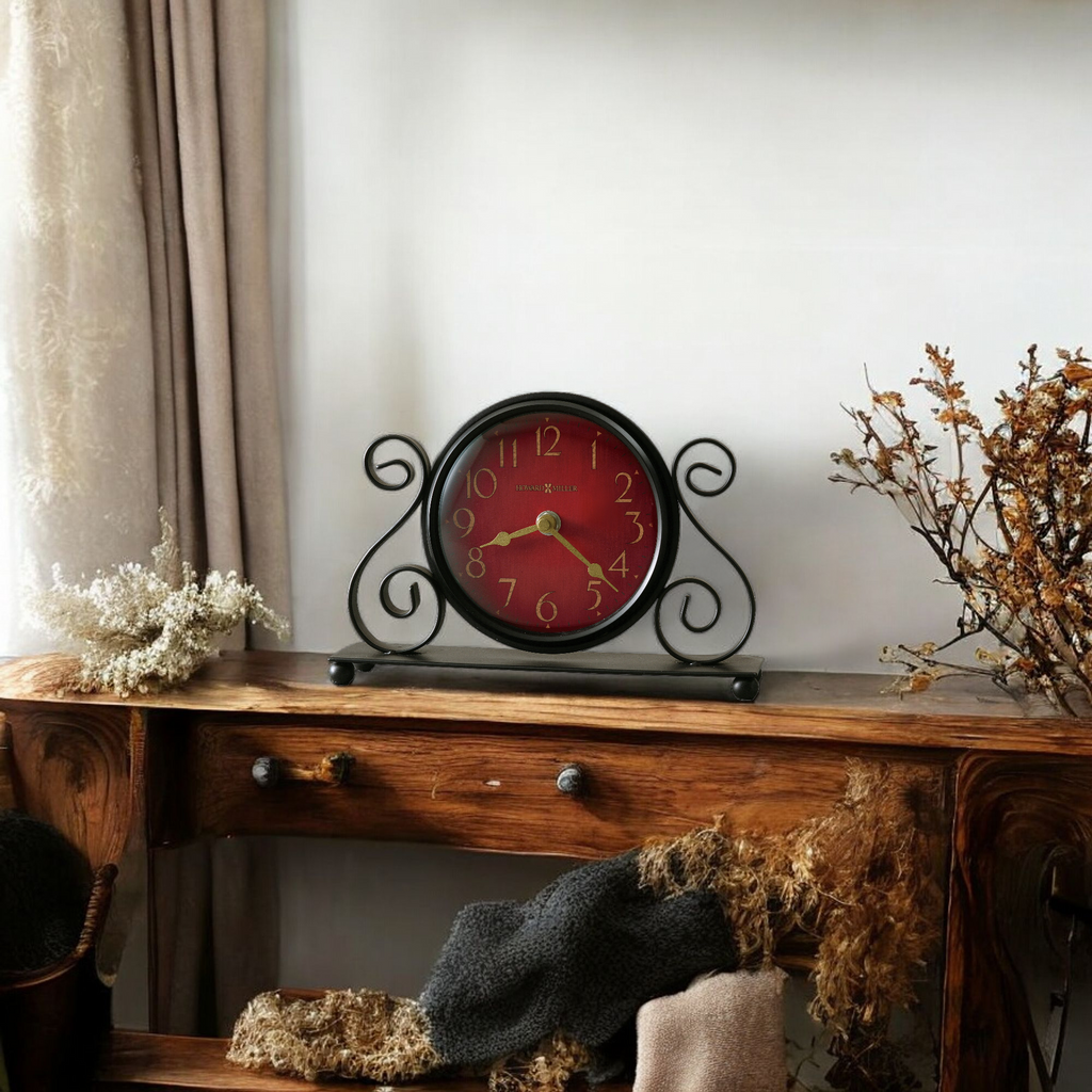 A decorative clock with a red face and black metal frame, placed on a wooden table with dried flowers and fabric.