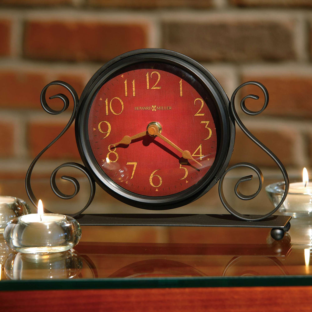 A decorative clock with a red face and ornate black stand, placed on a wooden surface next to candles.
