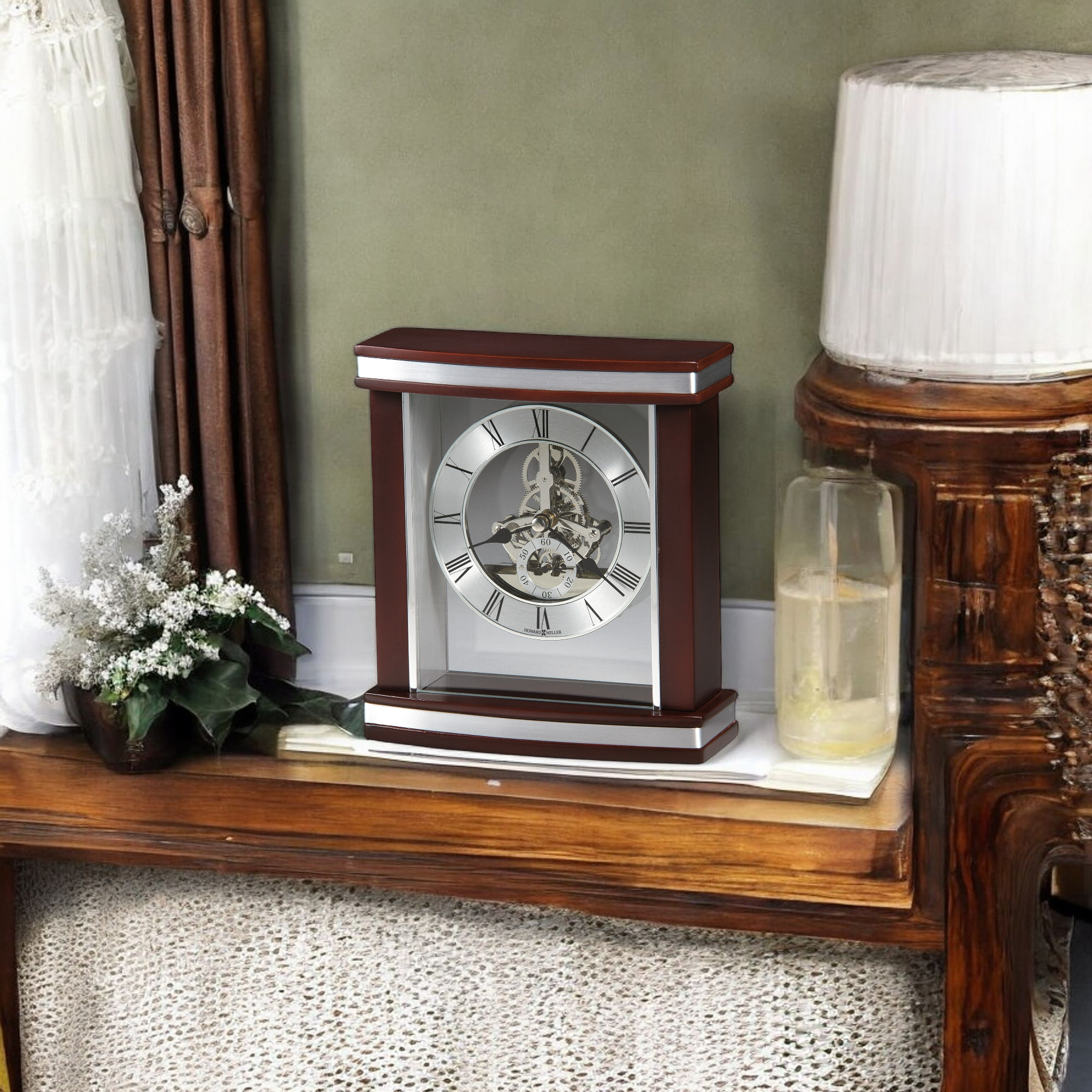 A decorative table clock on a wooden surface with a plant and a lamp.