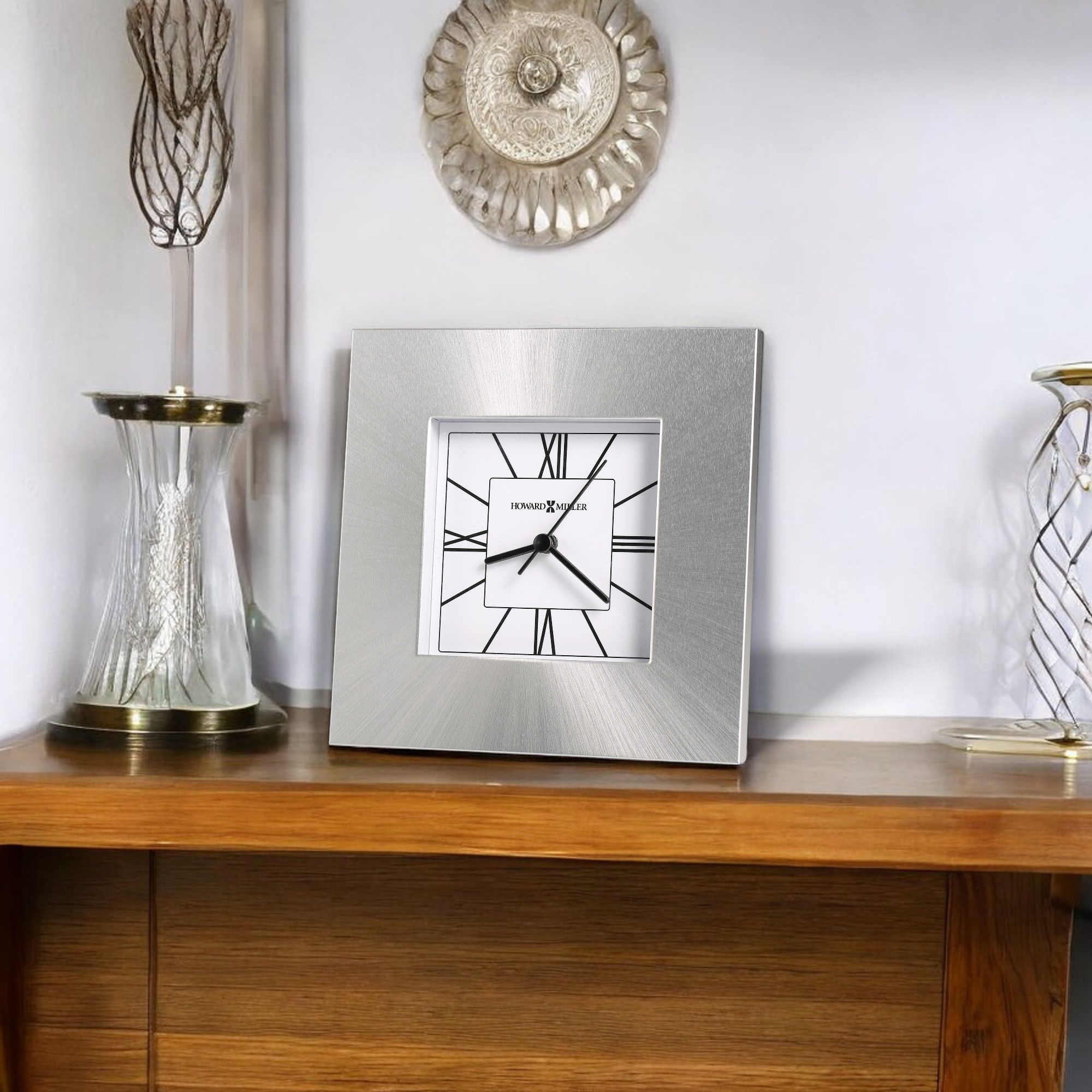 A silver-framed clock with Roman numerals displayed on a wooden shelf beside glass decorations.