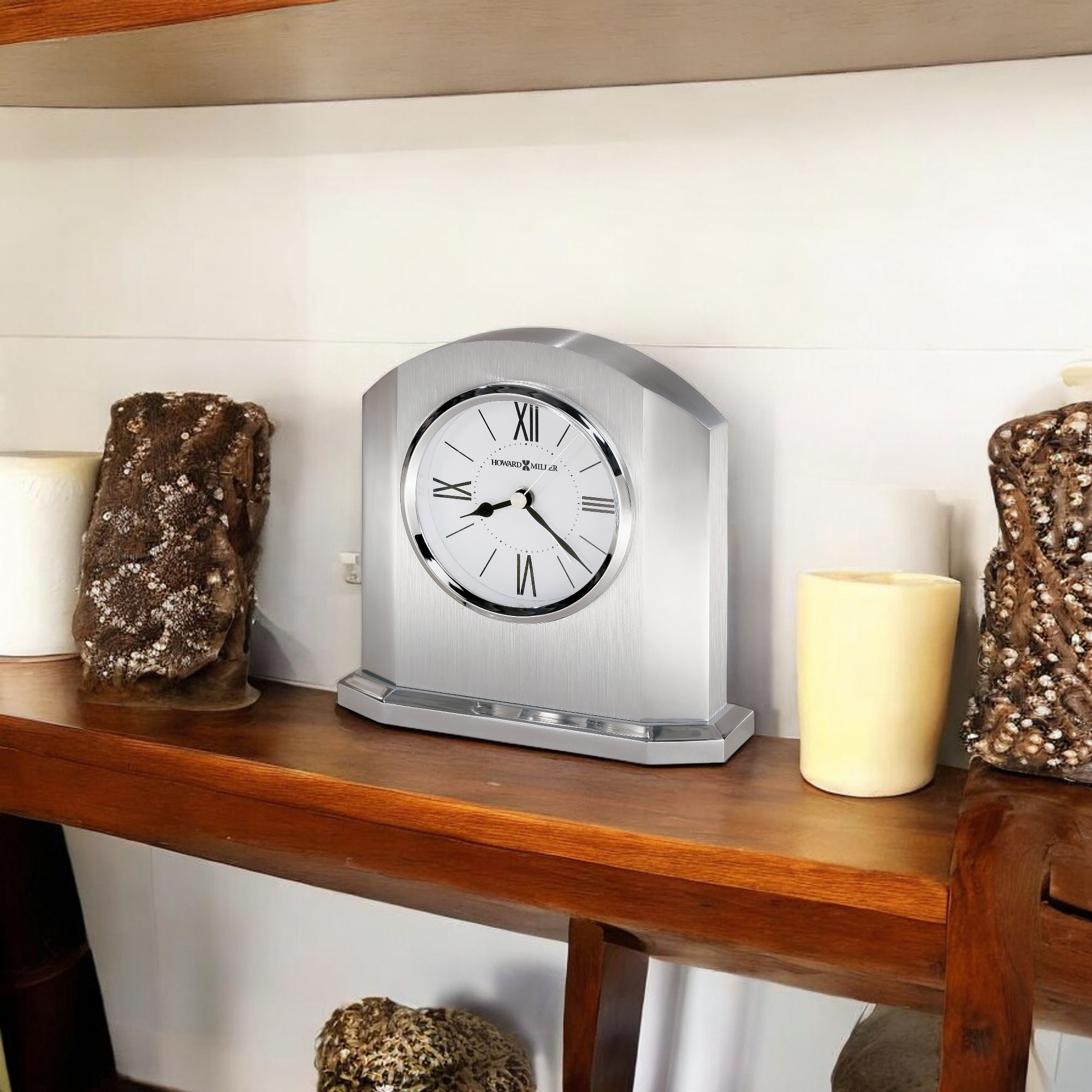 A silver wall clock placed on a wooden shelf decorated with candles and textured items.