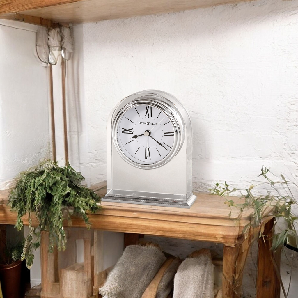A silver mantel clock with Roman numerals on a wooden shelf surrounded by greenery.