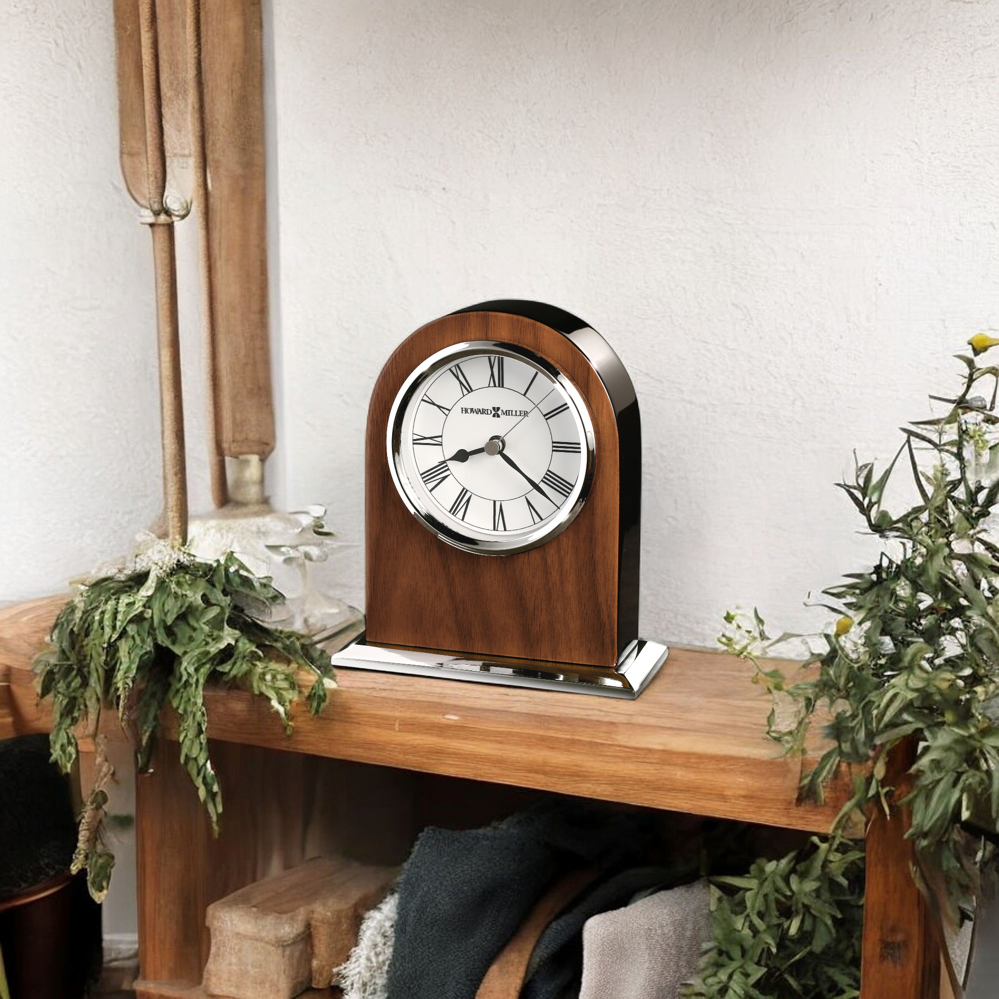 A classic wooden clock with a silver trim placed on a wooden shelf surrounded by plants.