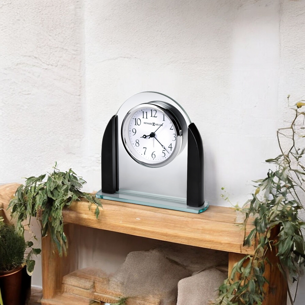 A modern clock with a sleek design on a wooden shelf, surrounded by plants.