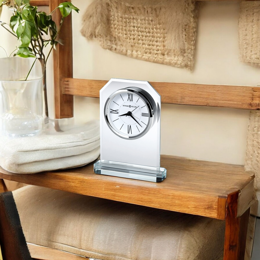 A stylish desk clock placed on a wooden shelf beside a folded blanket and a glass vase with plants.