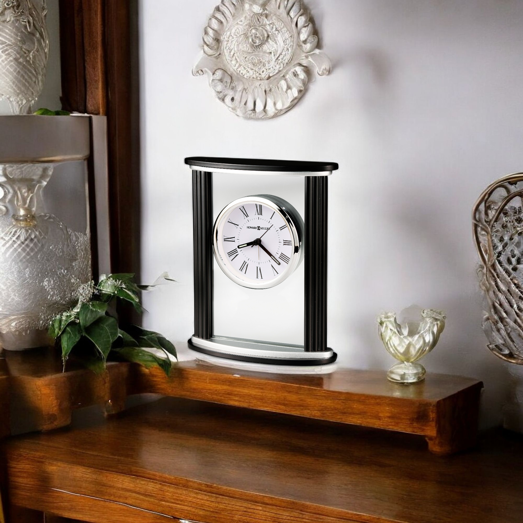 A decorative clock on a wooden shelf surrounded by glass vases and greenery.