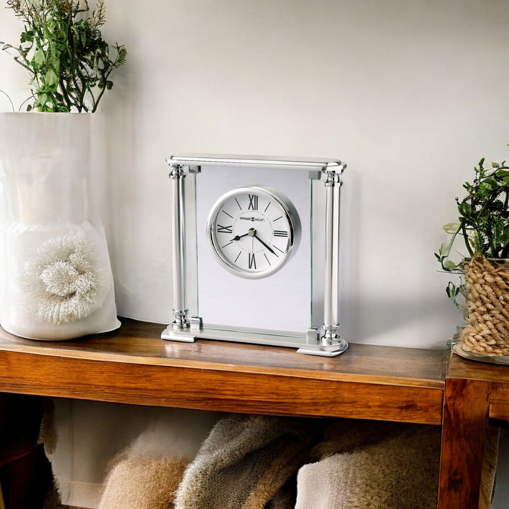 A decorative clock with a modern design displayed on a wooden shelf, surrounded by greenery and decorative items.