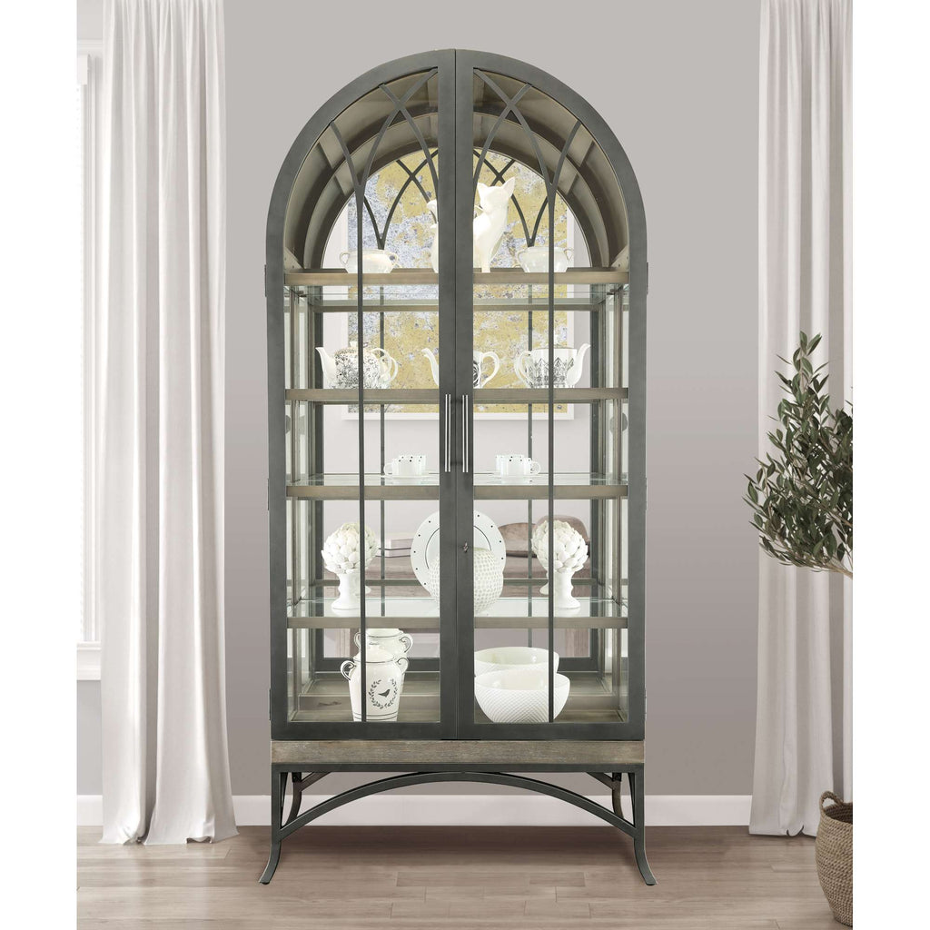 A stylish glass display cabinet with an arched top, showcasing decorative dishware against a neutral wall.