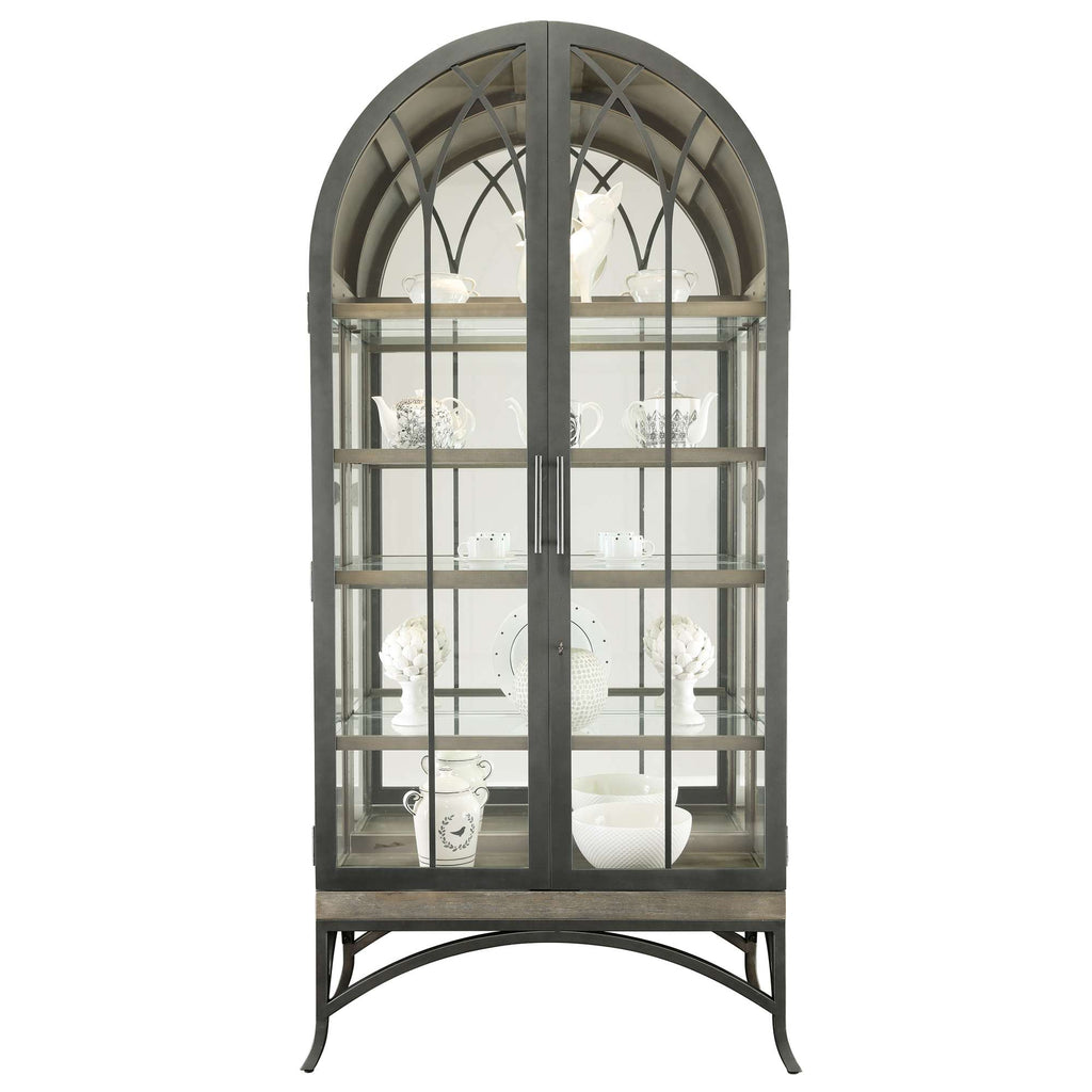A decorative glass display cabinet with an arched top, showcasing white ceramic items on wooden shelves.