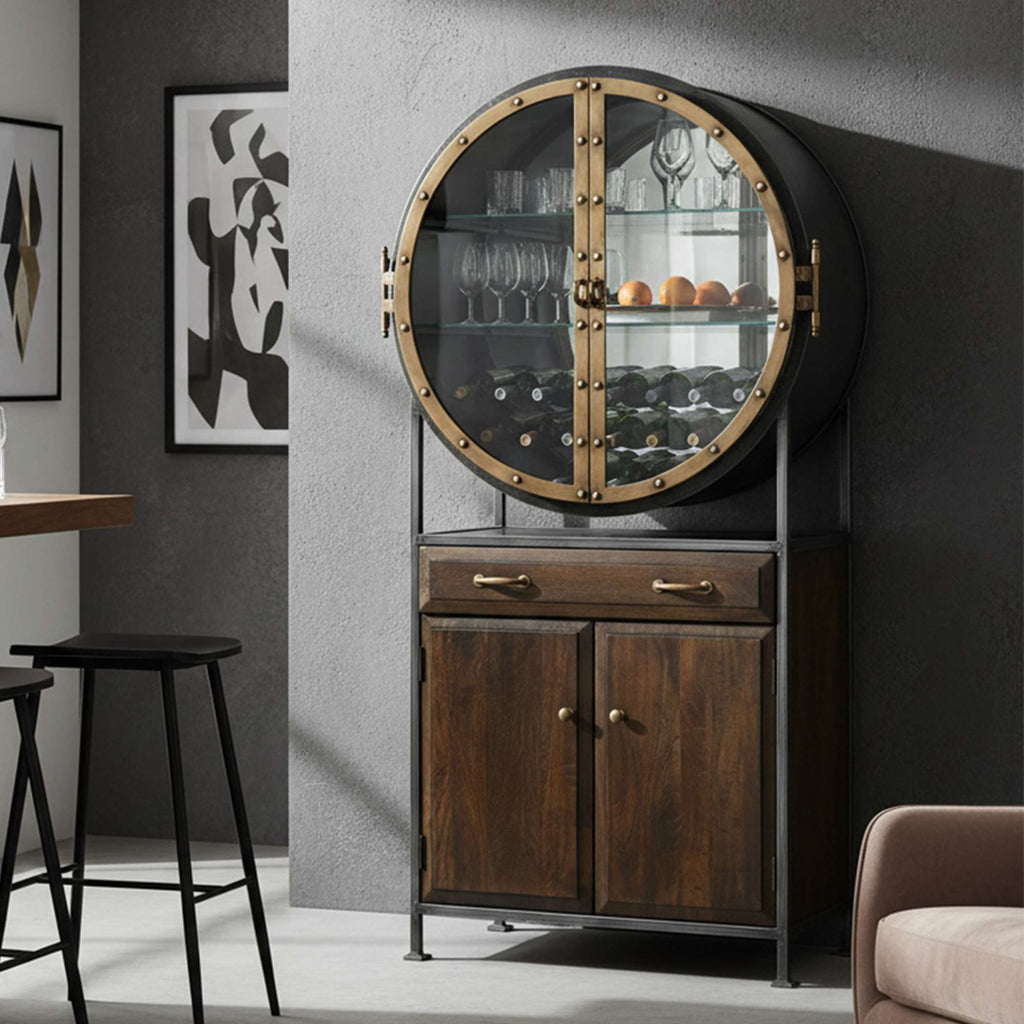 A stylish round glass cabinet with brass accents showcasing glasses and decor, set against a modern interior.
