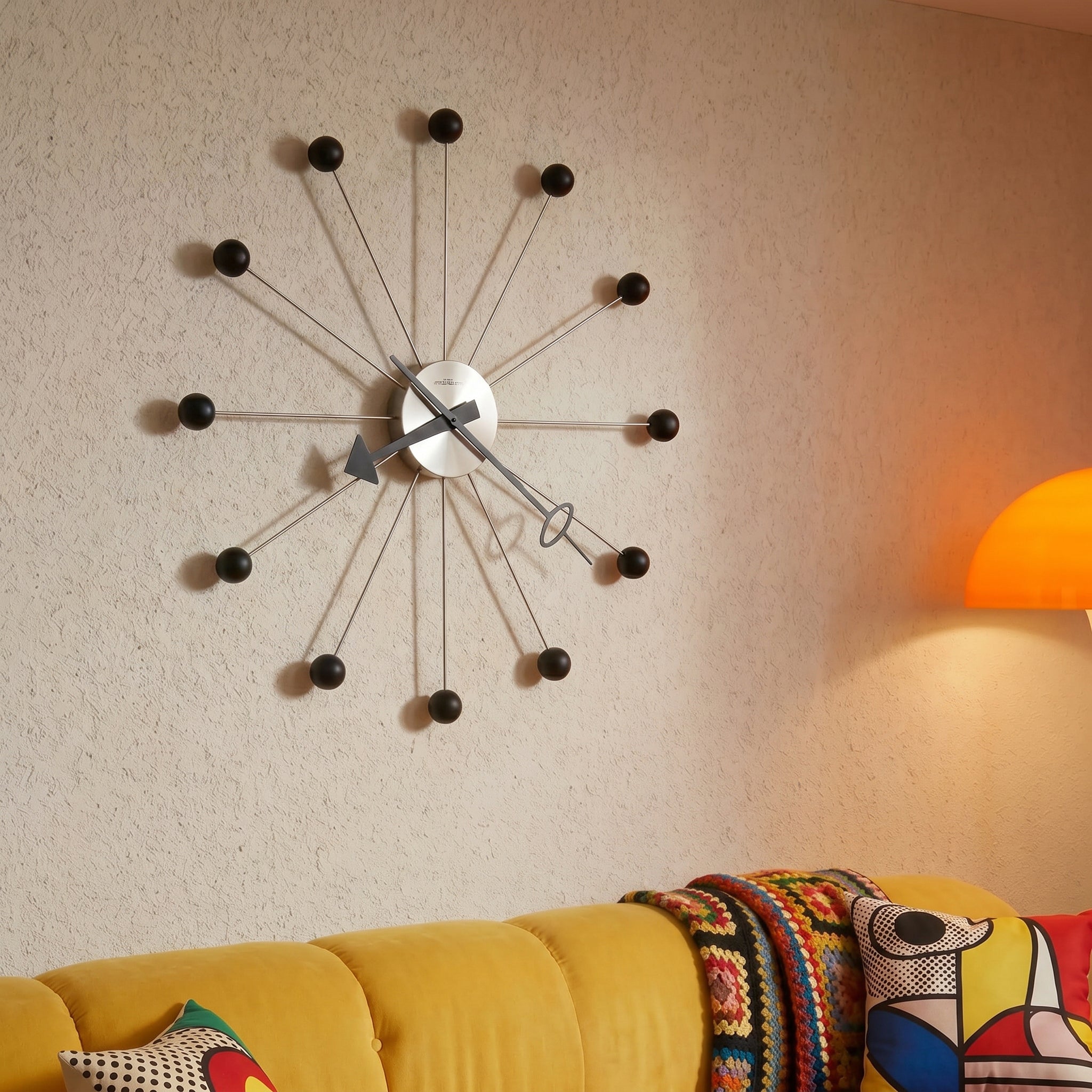 Modern wall clock on a beige wall with a yellow sofa and colorful cushions in the foreground.