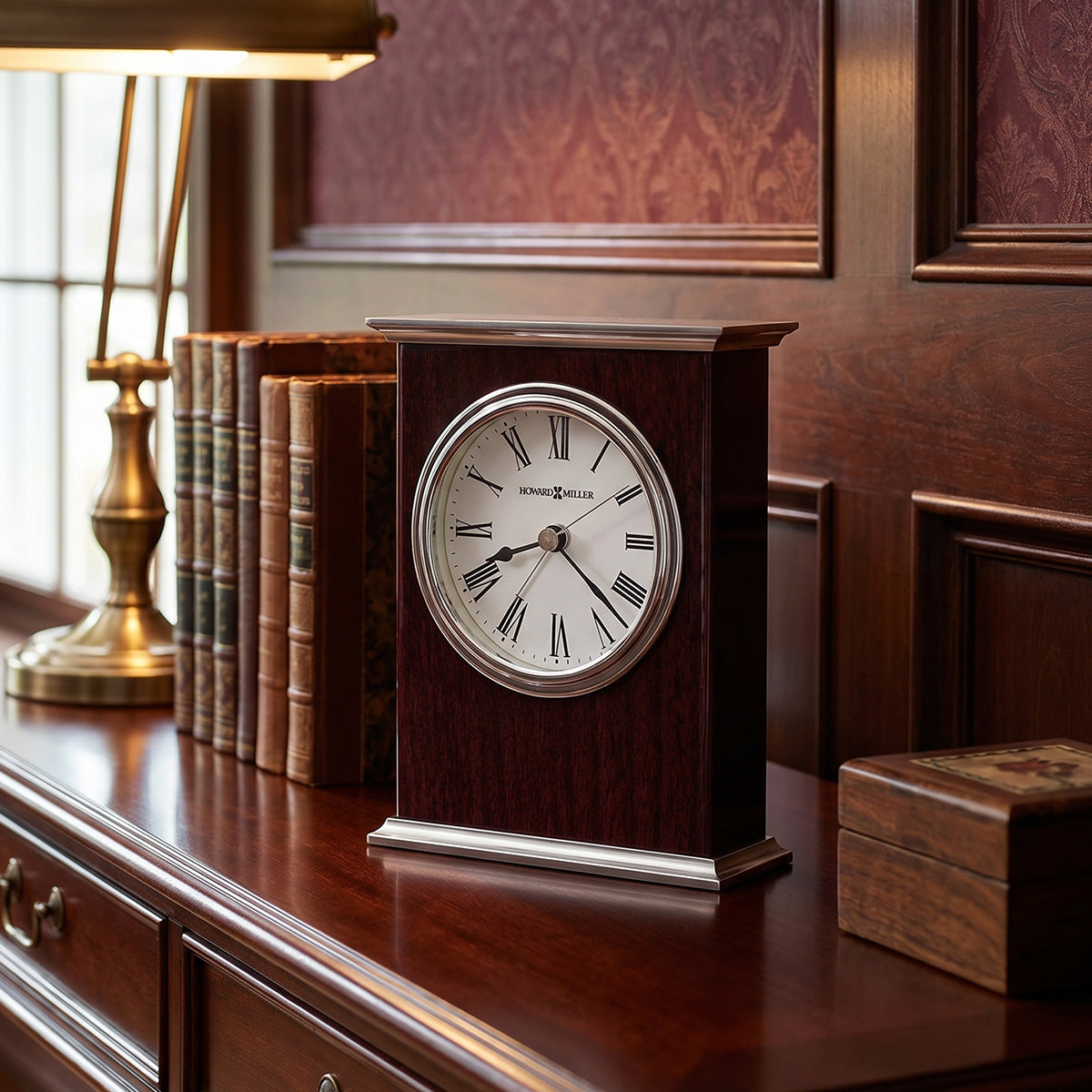 Wooden clock on a wooden surface with books and a lamp in the background