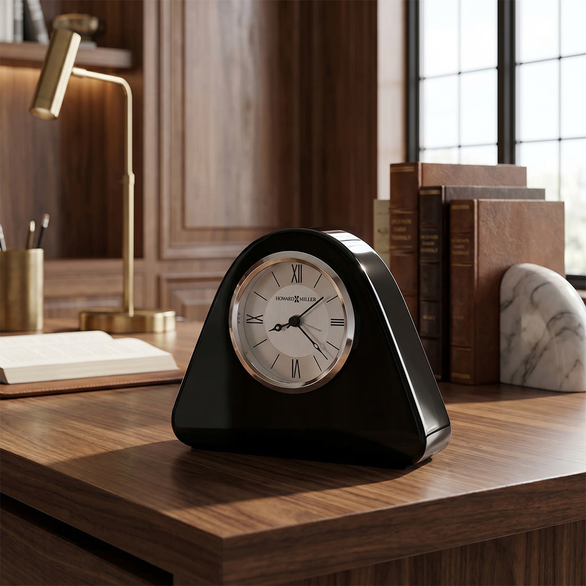 Black clock on a wooden desk with books and a lamp in the background
