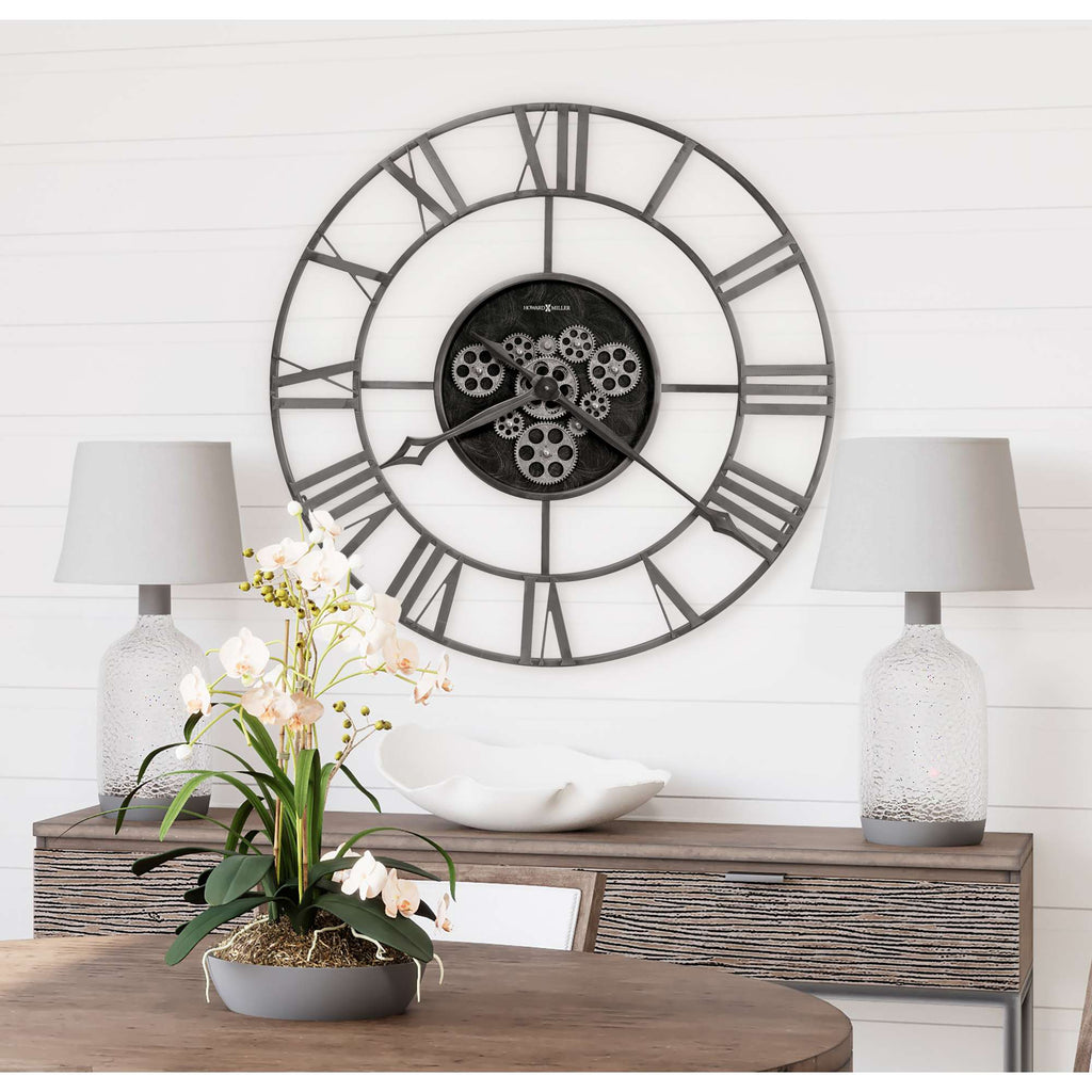 A stylish wall clock with a large circular frame and gears, displayed above a wooden table adorned with an orchid and two decorative lamps.
