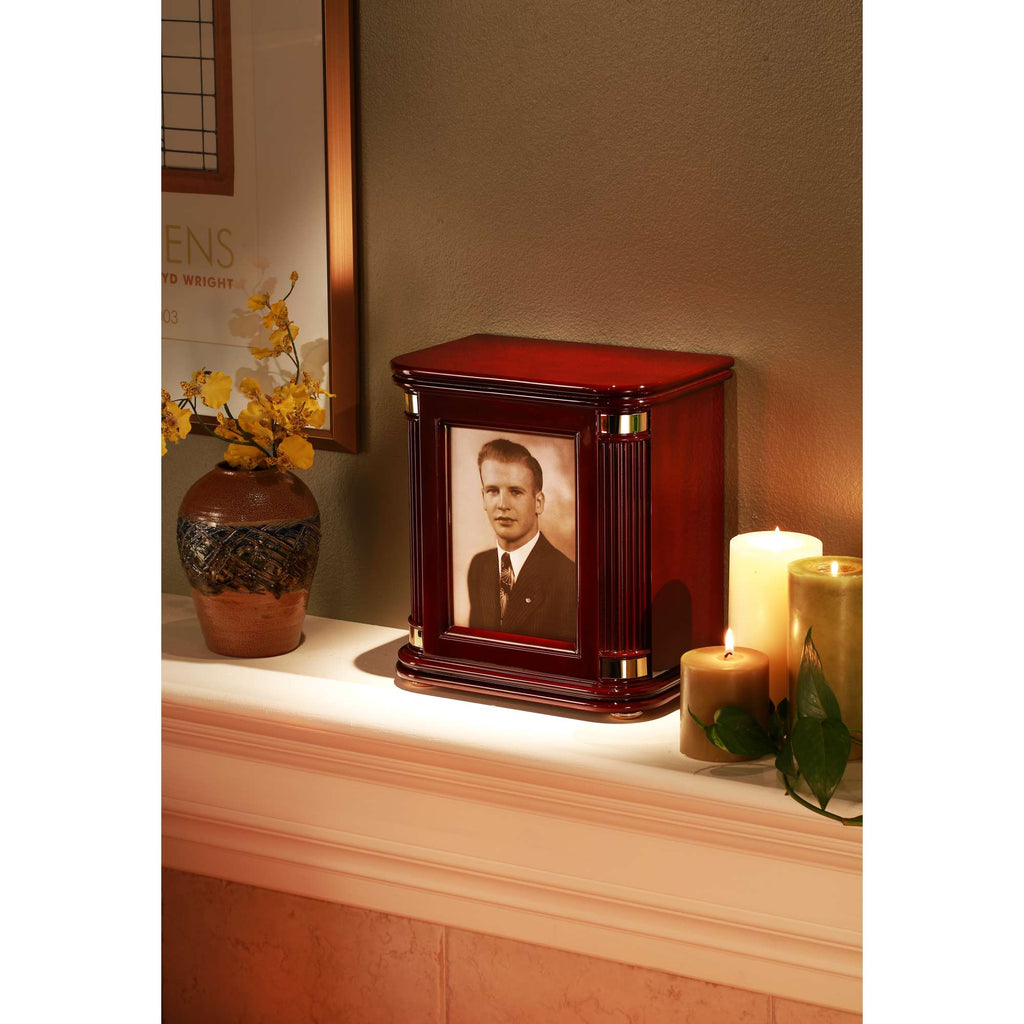 A decorative wooden urn with a framed photo on a mantel, surrounded by candles and a vase of flowers.