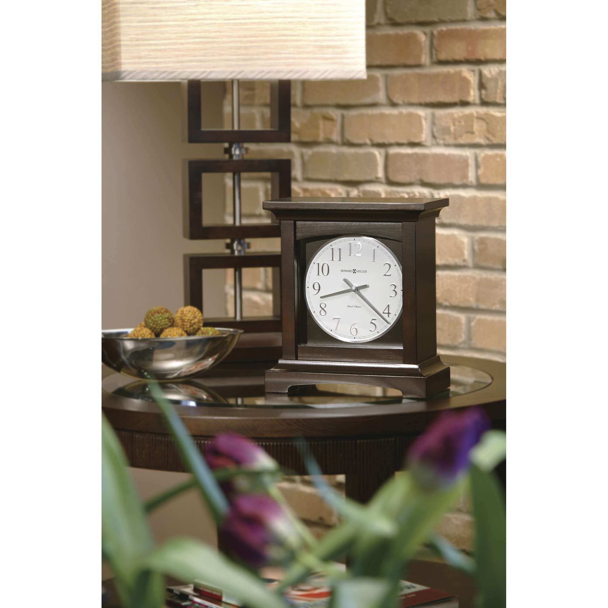 A wooden table clock next to a decorative bowl and a table lamp, with brick wall in the background.