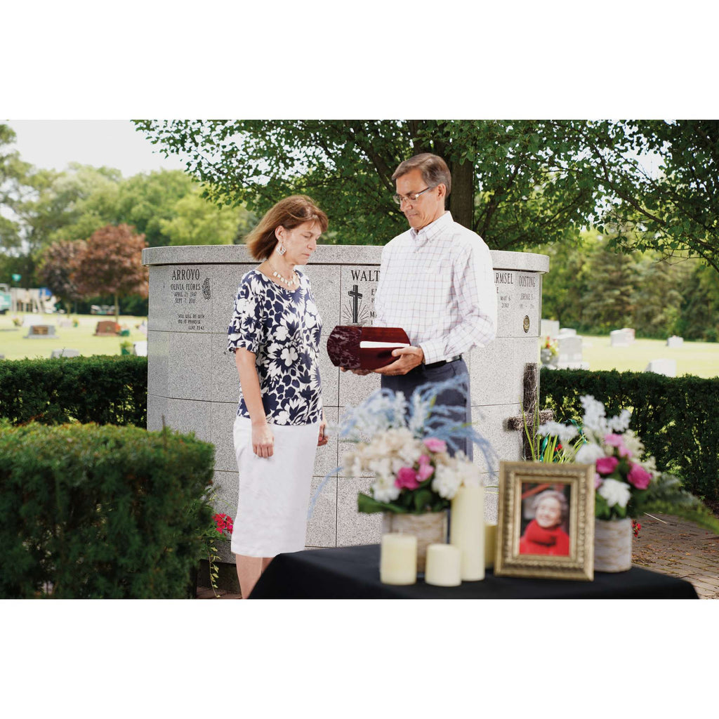 A man and woman standing by a memorial site, with flowers and photographs arranged on a table.