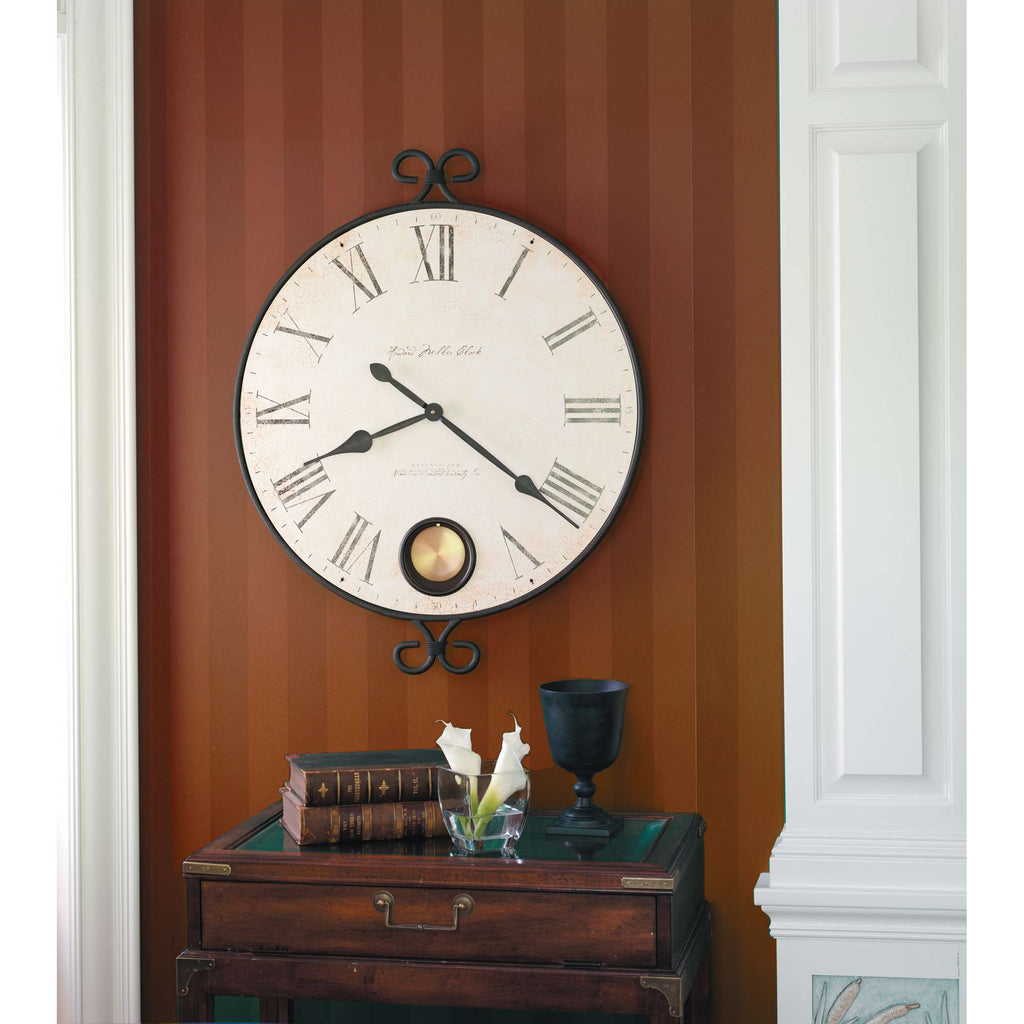 A classic wall clock with Roman numerals hangs above a wooden side table with books and a black vase containing white flowers against a striped orange wall.