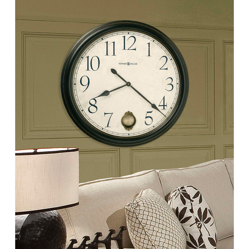 A large wall clock with a black frame, displaying classic numerals, hanging above a light-colored sofa with patterned cushions.