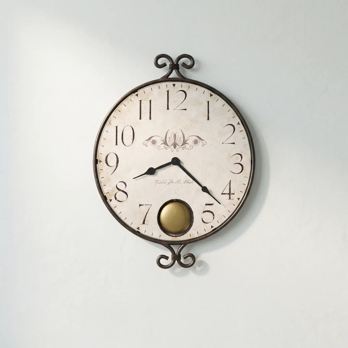 A round vintage-style wall clock with a distressed off-white face, black numerals and hands, and an ornate dark metal frame with scroll details at the top and bottom. A small gold-toned circular accent is visible near the lower center.