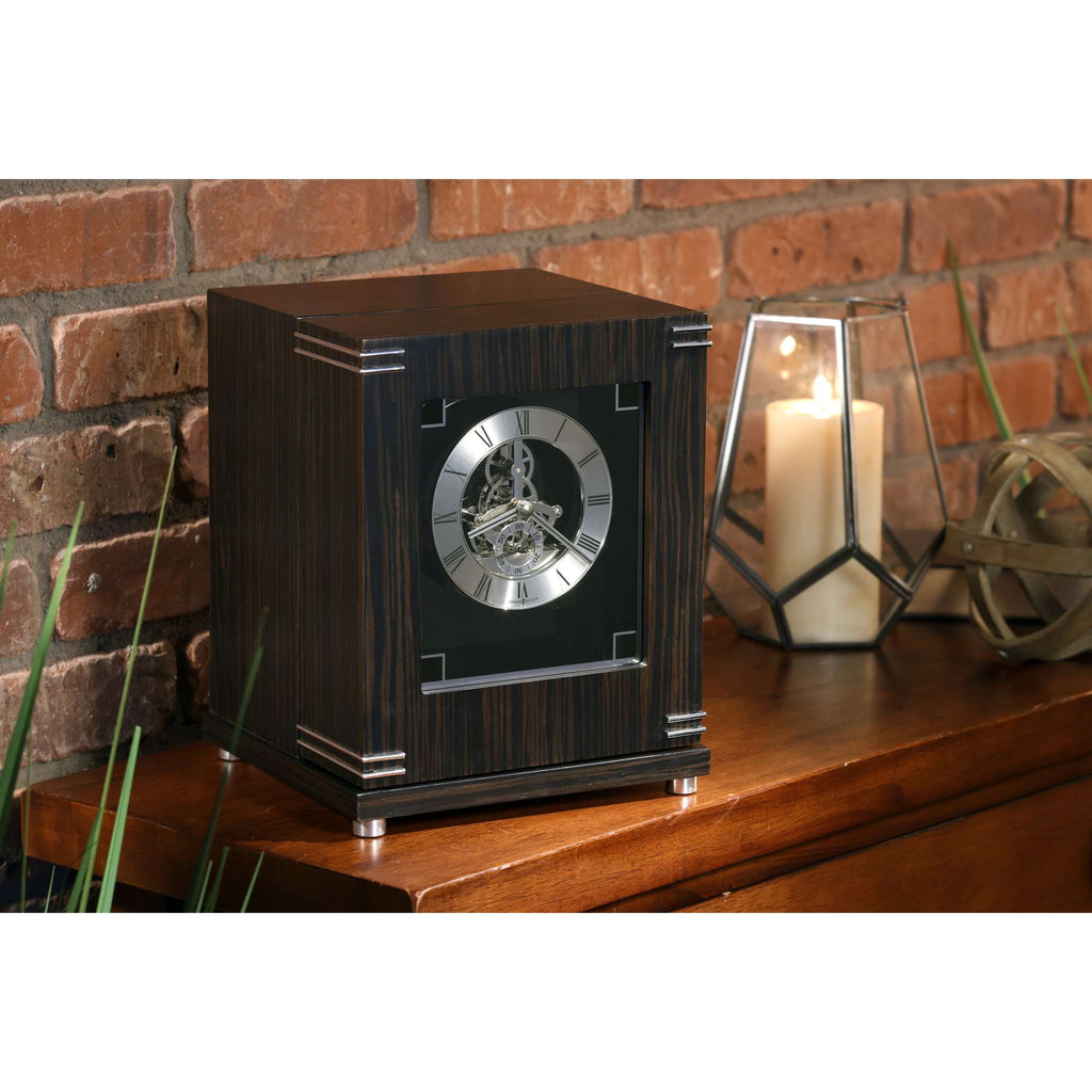 A stylish wooden clock on a table against a brick wall, with candles and decorative items nearby.