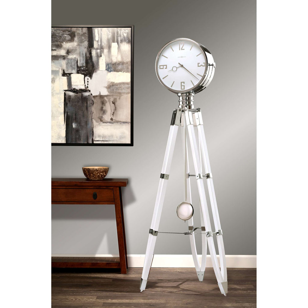 A modern standing clock with a sleek design on a tripod base, next to a wall art piece.