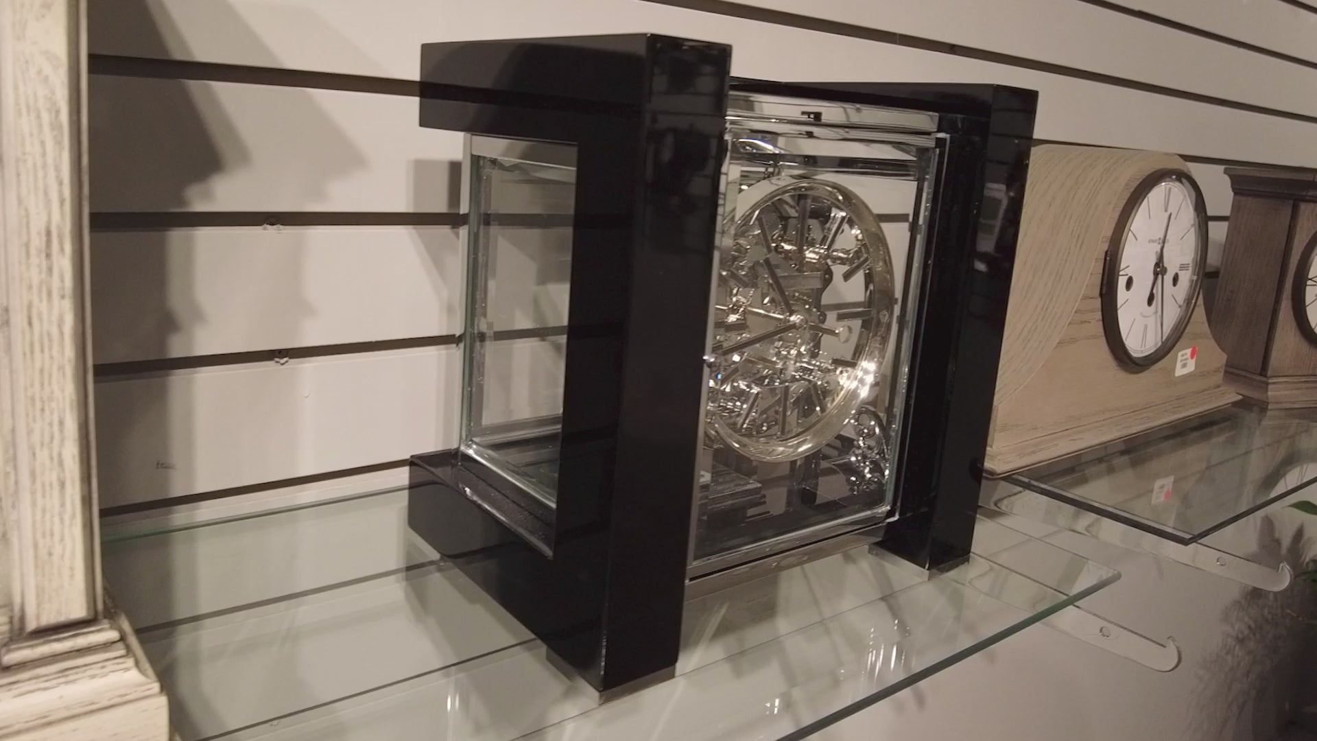A modern black clock with a transparent casing displayed on a glass shelf.