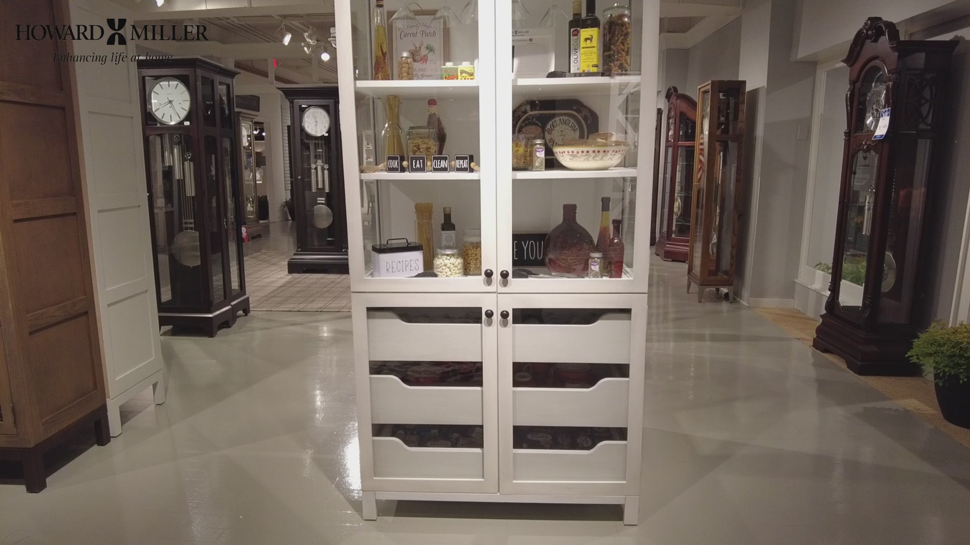 A white display cabinet with glass shelves and drawers, surrounded by clock furniture in a showroom.