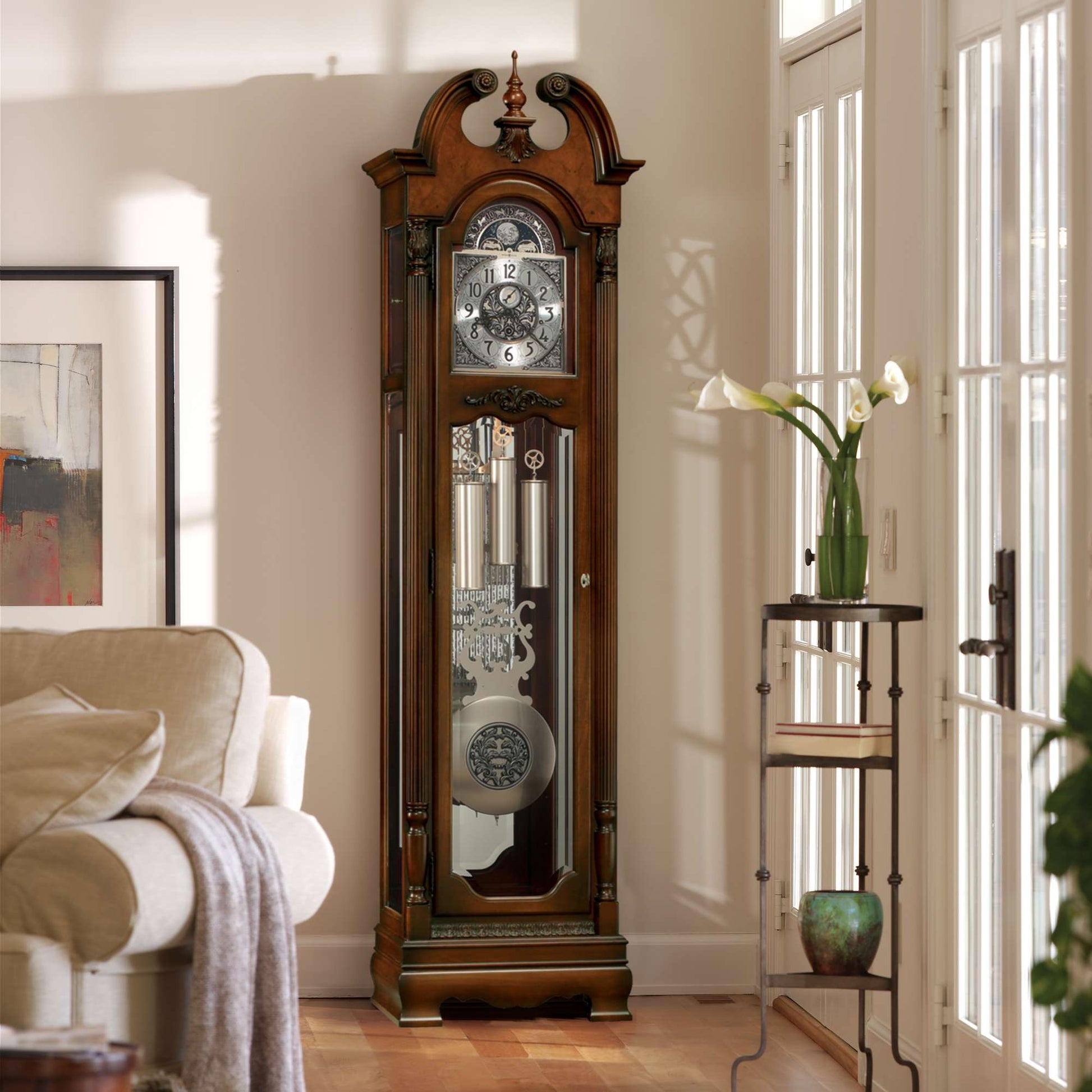 A tall, ornate grandfather clock next to a couch and a decorative plant stand in a well-lit room.