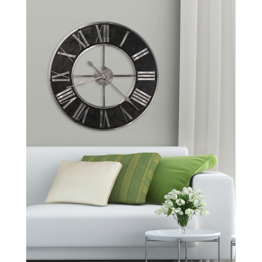 A modern wall clock with Roman numerals hanging above a white couch adorned with green and cream cushions.