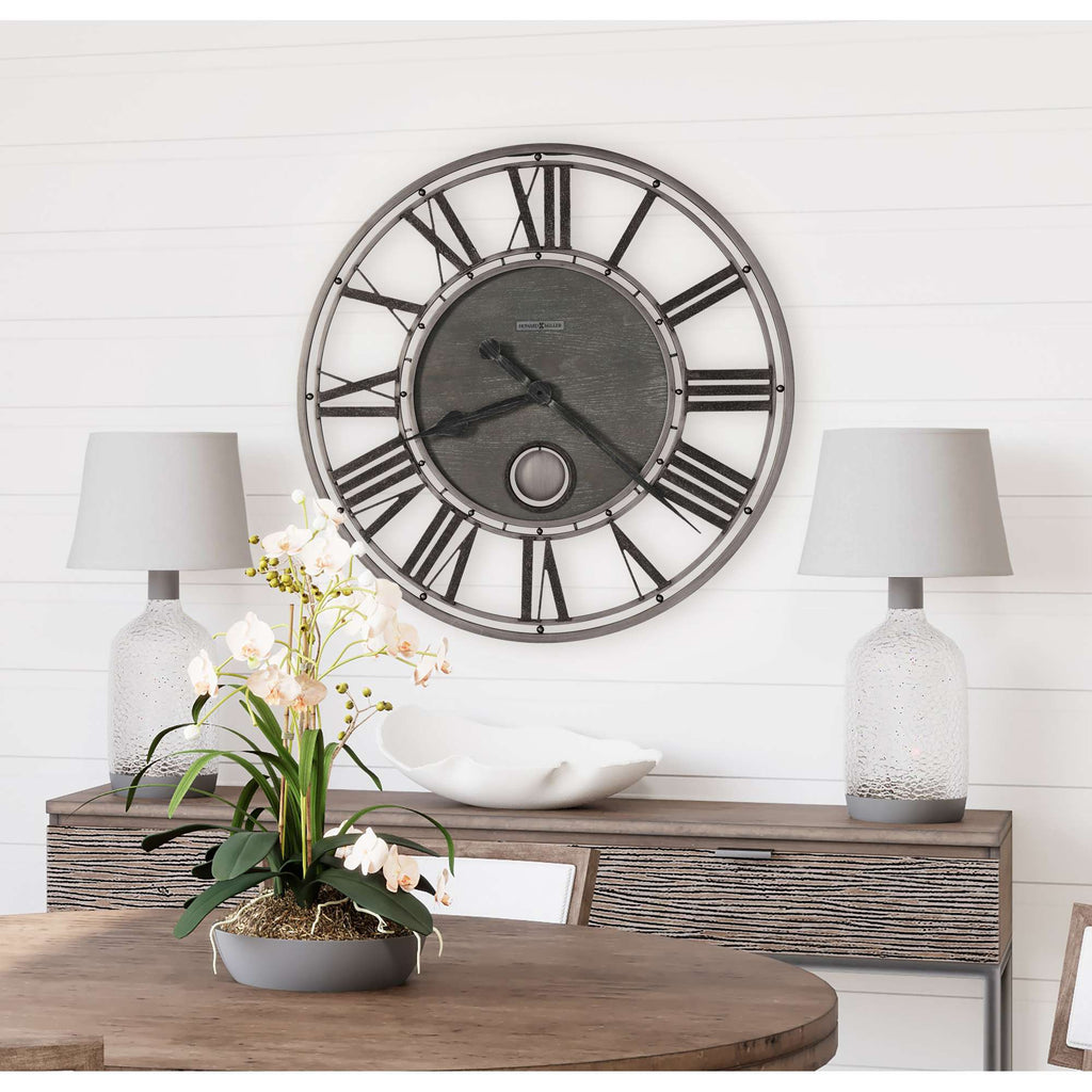 A modern wall clock with Roman numerals above a wooden table adorned with a potted plant and two table lamps.