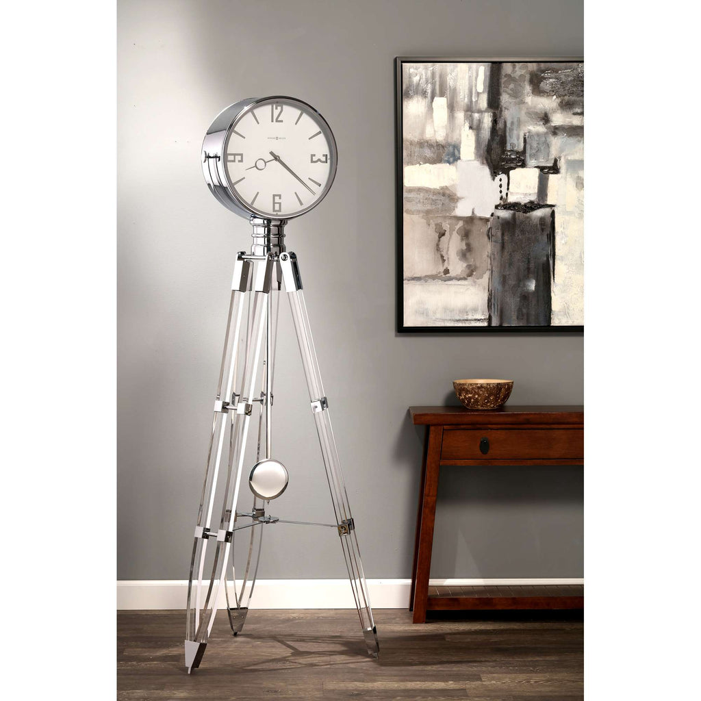 A modern floor clock with a silver finish and tripod legs, displayed beside an abstract artwork and a wooden table.